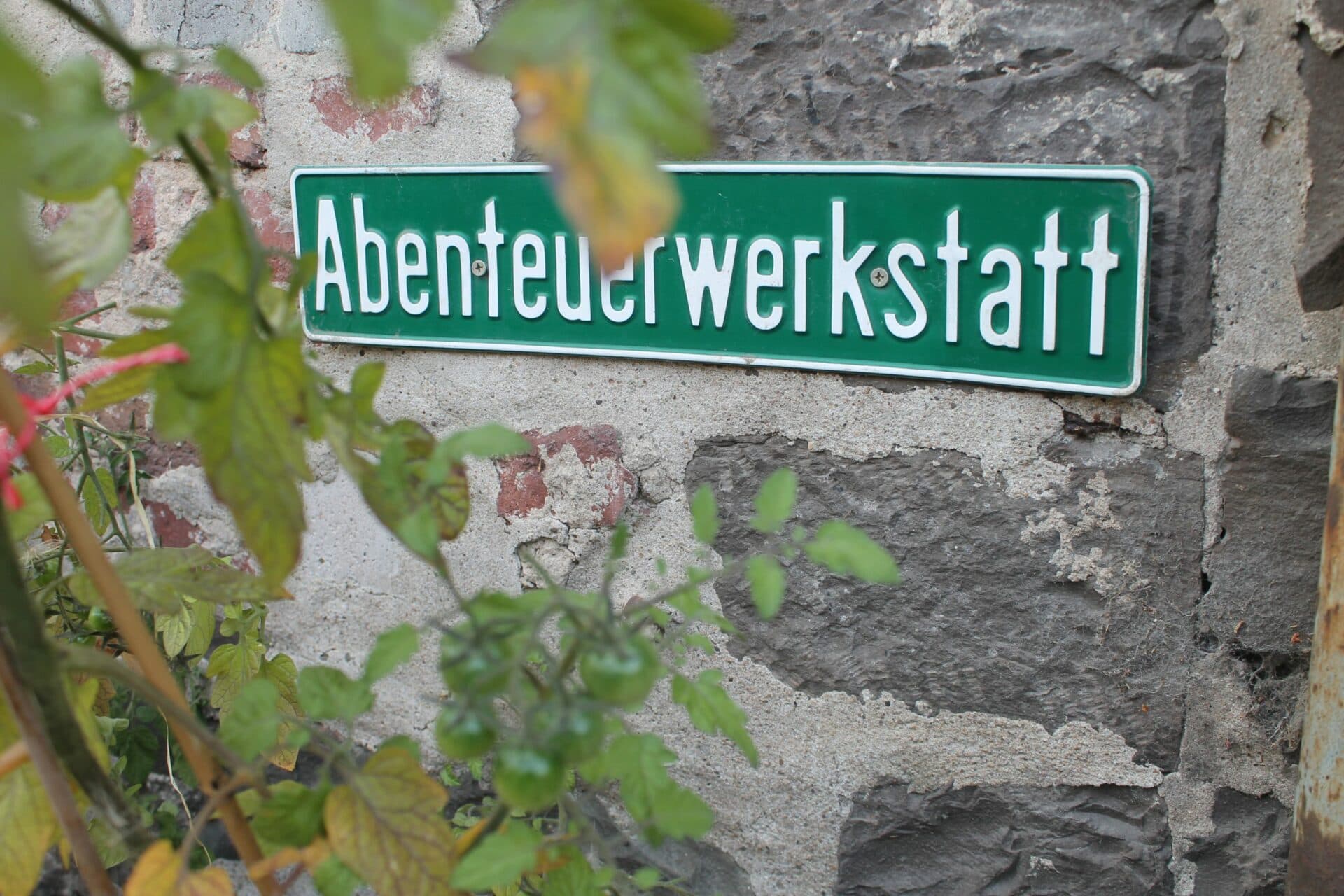 Grünes Straßenschild mit der Aufschrift „Abenteuerwerkstatt“ an einer alten Bruchsteinmauer, davor Tomatenpflanzen
